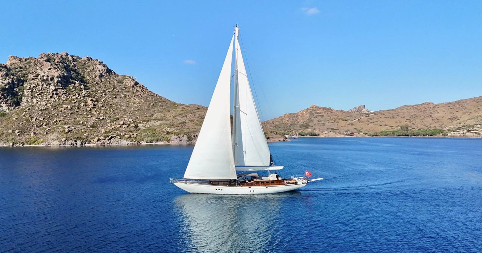 A stunning sailing yacht glides gracefully through calm, blue waters against a backdrop of rugged coastal hills. aboard PORTOBELLO Yacht for Sale