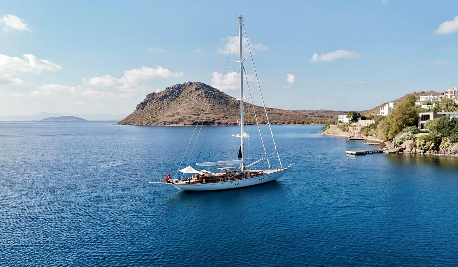 A sleek sailing yacht is anchored in a serene bay with a picturesque backdrop of hills and clear blue water. aboard PORTOBELLO Yacht for Sale