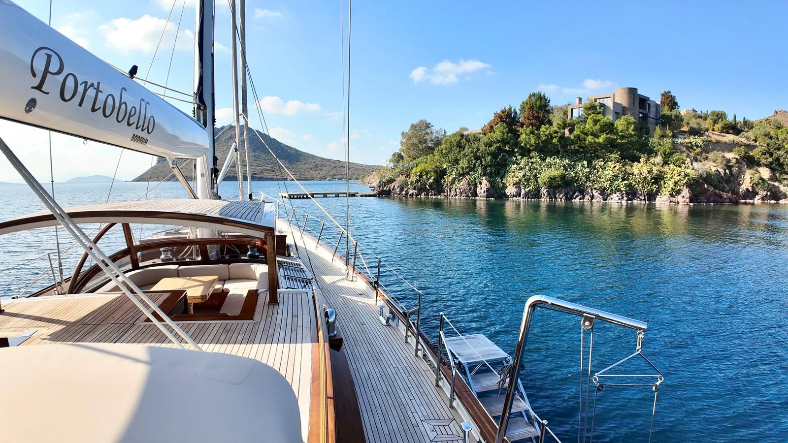 A stunning yacht is anchored in a serene bay with lush greenery and a modern villa in the background. aboard PORTOBELLO Yacht for Sale