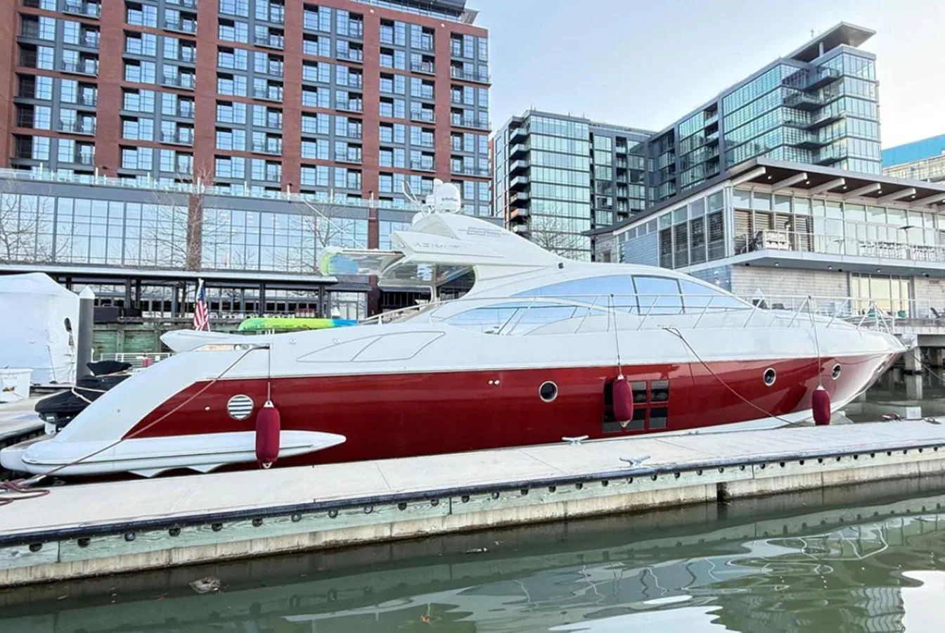 A sleek yacht with a red hull is docked in a modern marina with urban buildings in the background. aboard 2007 AZIMUT 68S Yacht for Sale