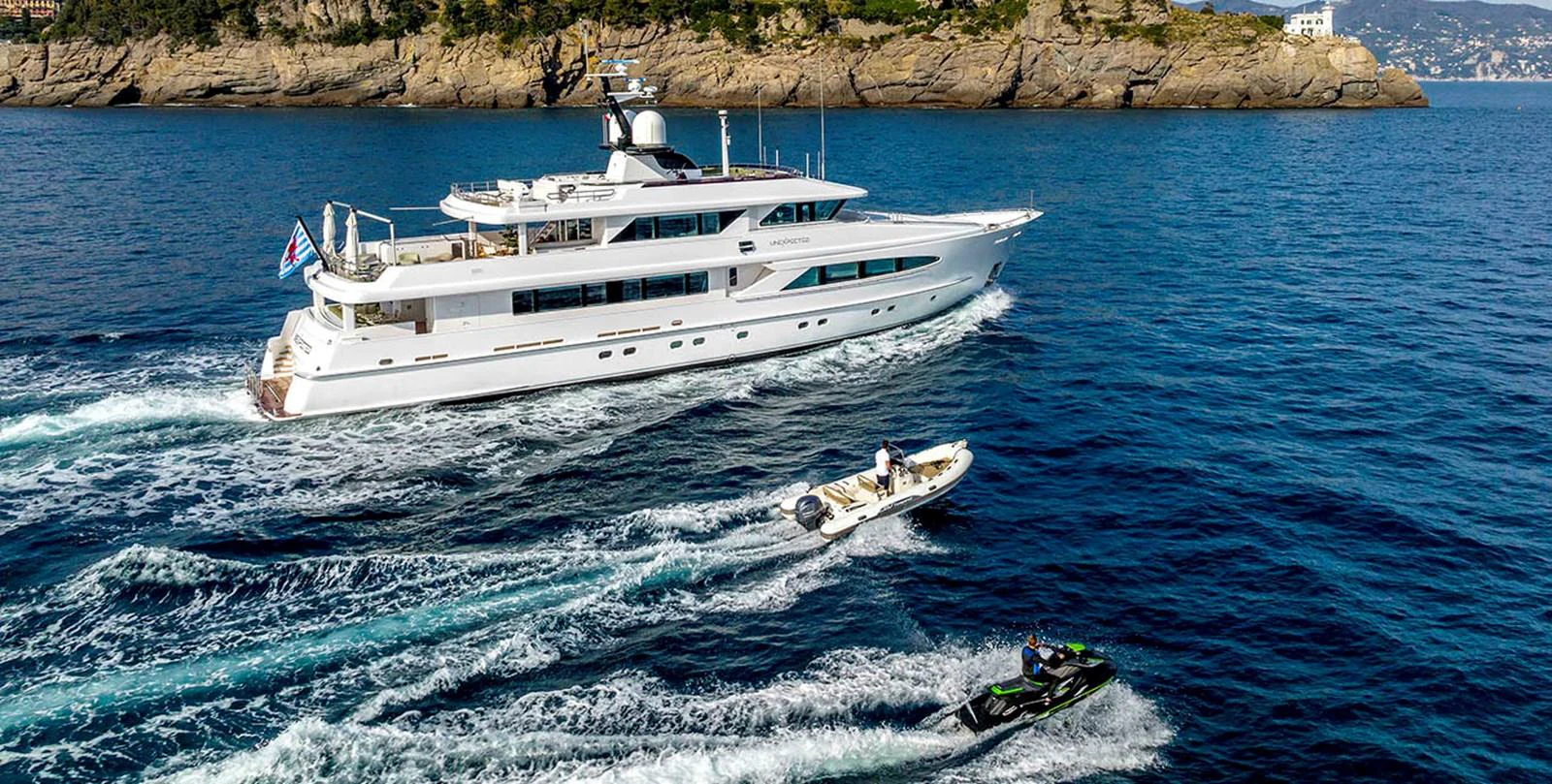 A luxurious yacht cruises along the coastline, accompanied by a tender and a jet ski. aboard UNEXPECTED Yacht for Sale
