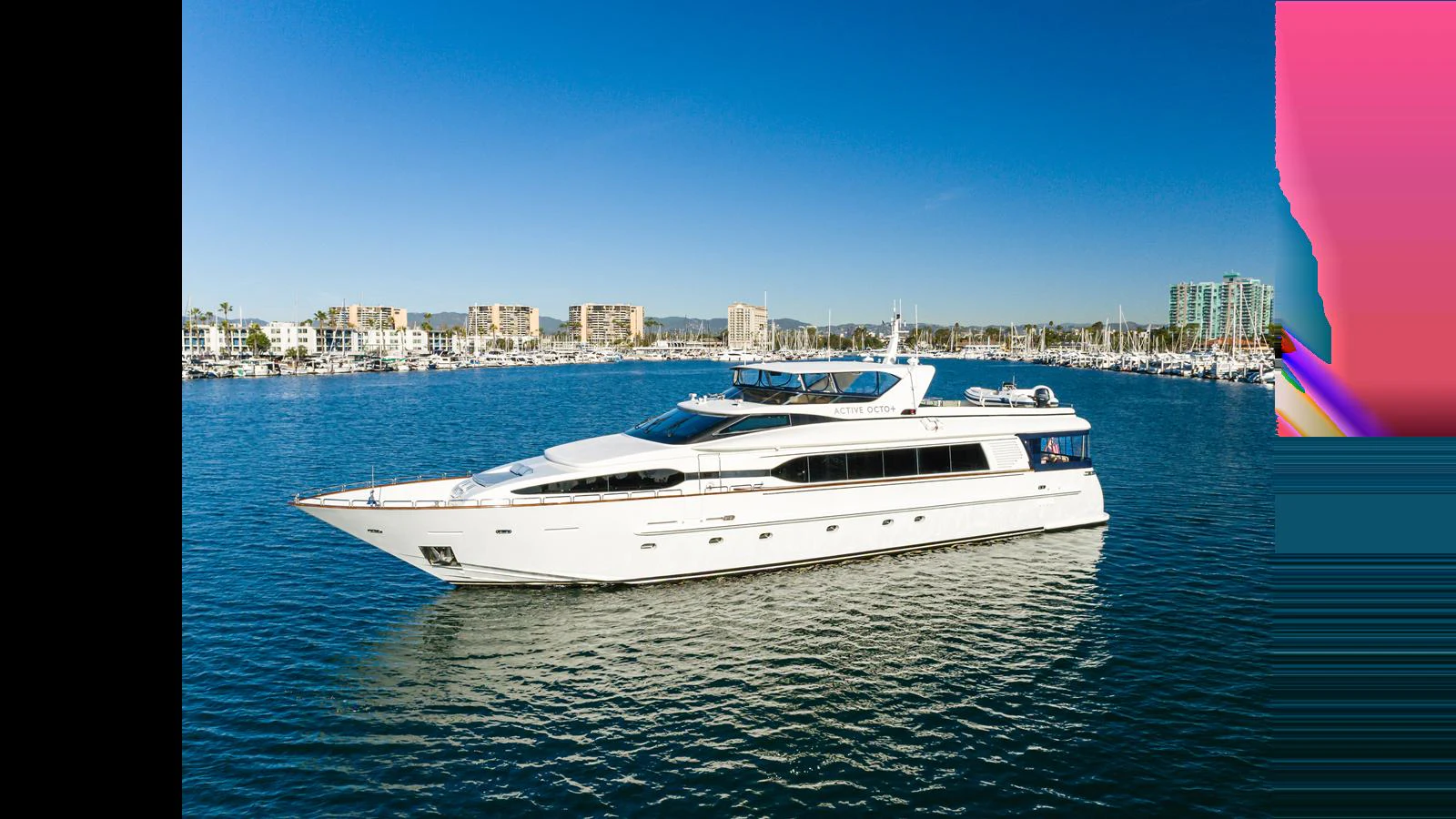 A sleek white yacht cruises gracefully on calm blue waters with a marina and cityscape in the background. aboard ACTIVE OCTO Yacht for Sale