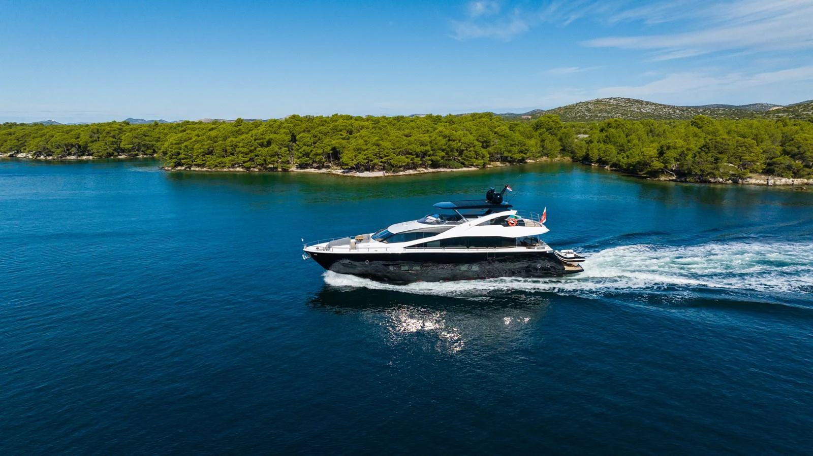 A sleek yacht cruises through calm, clear waters with a lush, green shoreline in the background. aboard BLACK MAMBA Yacht for Sale