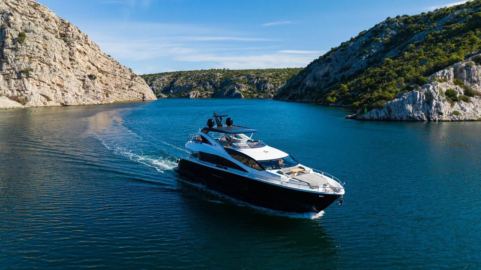 A sleek yacht cruises through a serene, rocky inlet under a clear blue sky. aboard BLACK MAMBA Yacht for Sale