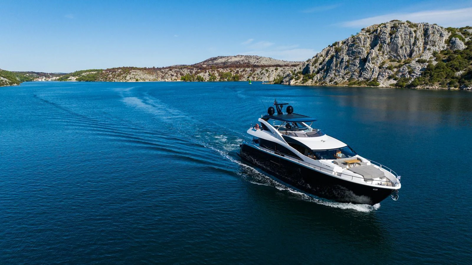 A sleek yacht cruises through a serene, rocky coastal landscape under a clear blue sky. aboard BLACK MAMBA Yacht for Sale