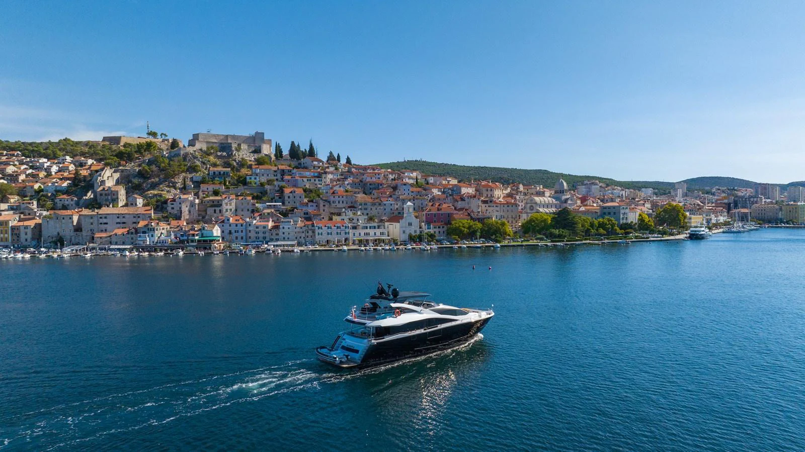A sleek yacht cruises along a picturesque coastal town under a clear blue sky. aboard BLACK MAMBA Yacht for Sale