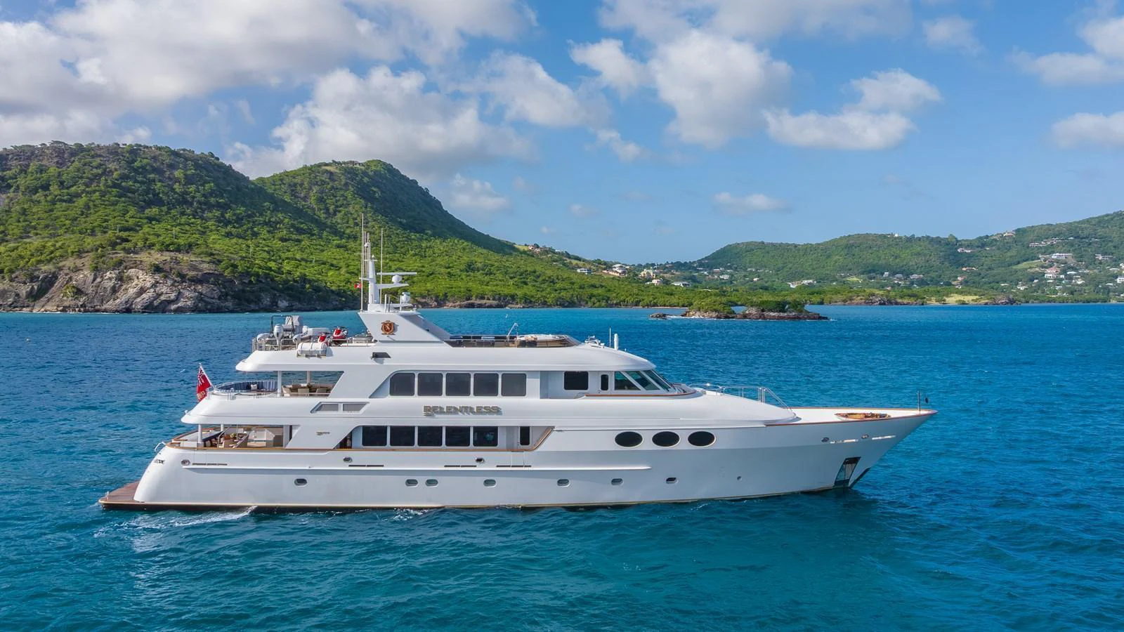 A luxurious yacht cruising through clear blue waters with scenic hills in the background. aboard RELENTLESS Yacht for Sale