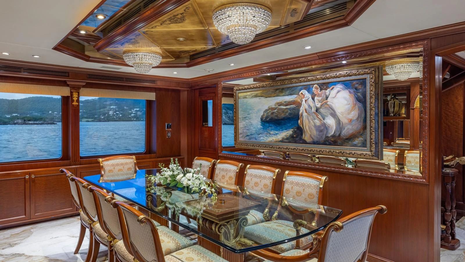 The yacht's dining area features an elegant glass table, luxurious seating, and opulent decor with a large painting and chandeliers. aboard RELENTLESS Yacht for Sale