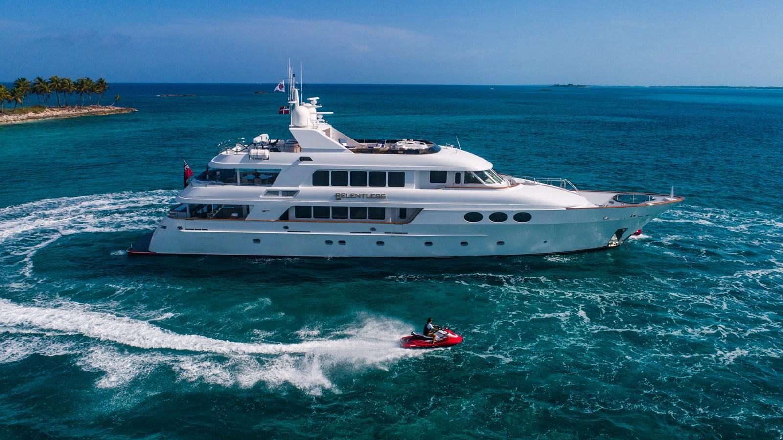 A luxurious yacht cruises through clear blue waters, accompanied by a jet ski. aboard RELENTLESS Yacht for Sale