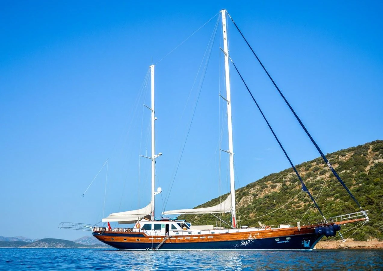 A stunning sailing yacht with twin masts glides gracefully on calm waters against a backdrop of lush hills. aboard PAPA JOE Yacht for Sale