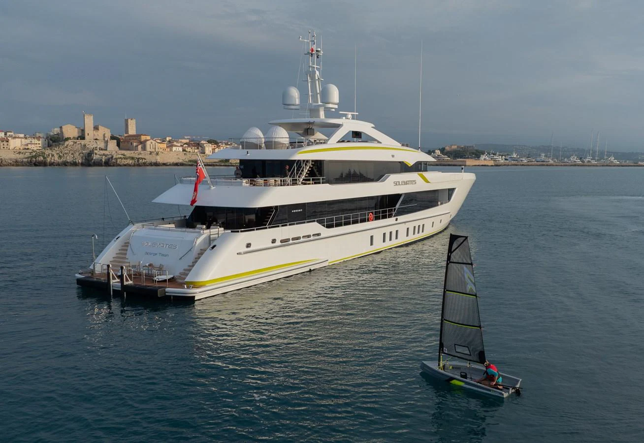 A sleek, modern yacht is anchored near a coastal town, with a small sailboat nearby. aboard SOLEMATES Yacht for Sale