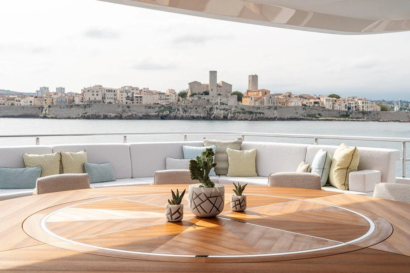 The aft deck features a stylish circular wooden table with potted plants, surrounded by comfortable seating and offering a scenic view of the coastline. aboard SOLEMATES Yacht for Sale