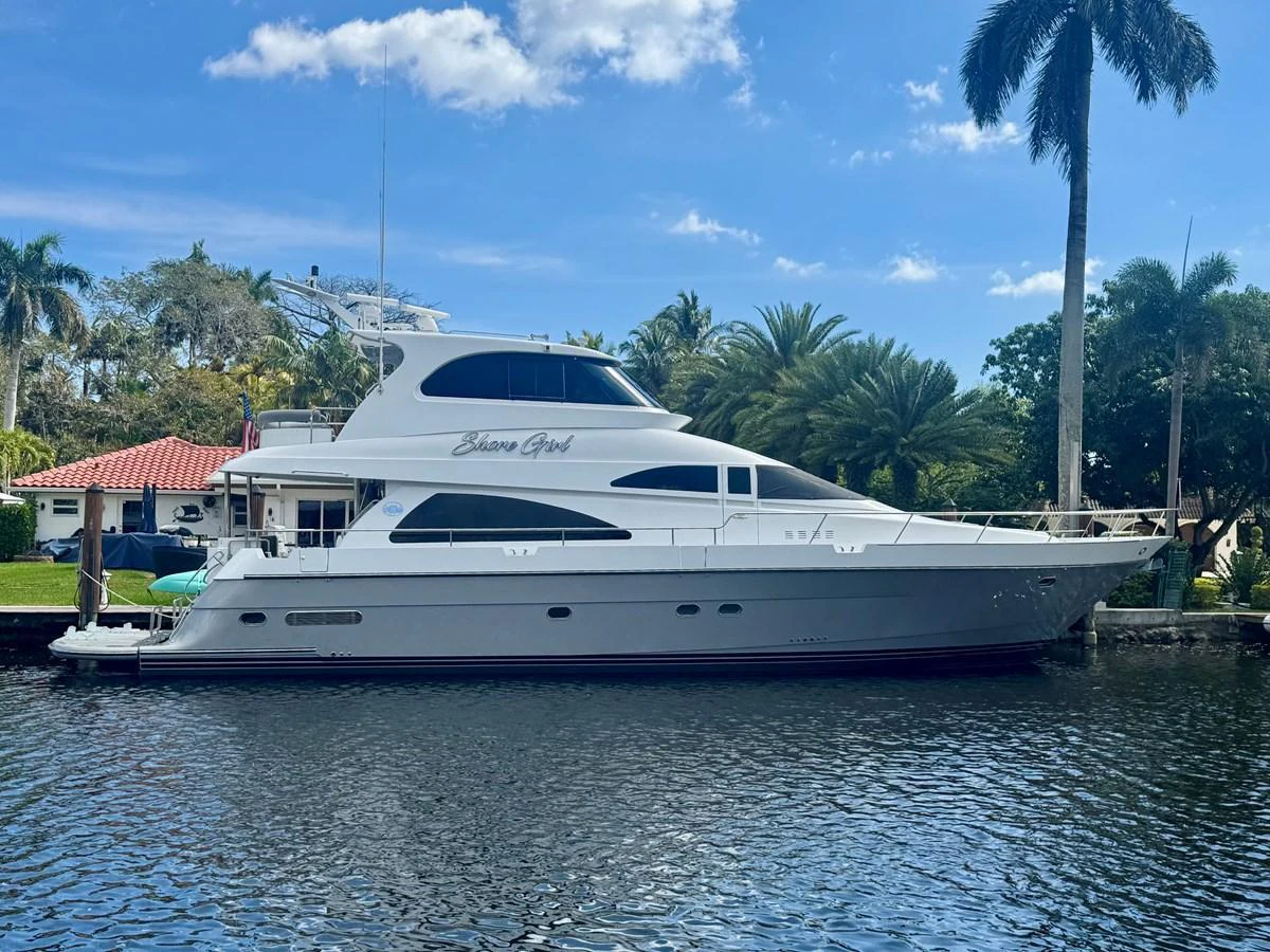 A sleek yacht is docked in a serene waterfront setting under a clear blue sky. aboard SHORE GIRL Yacht for Sale