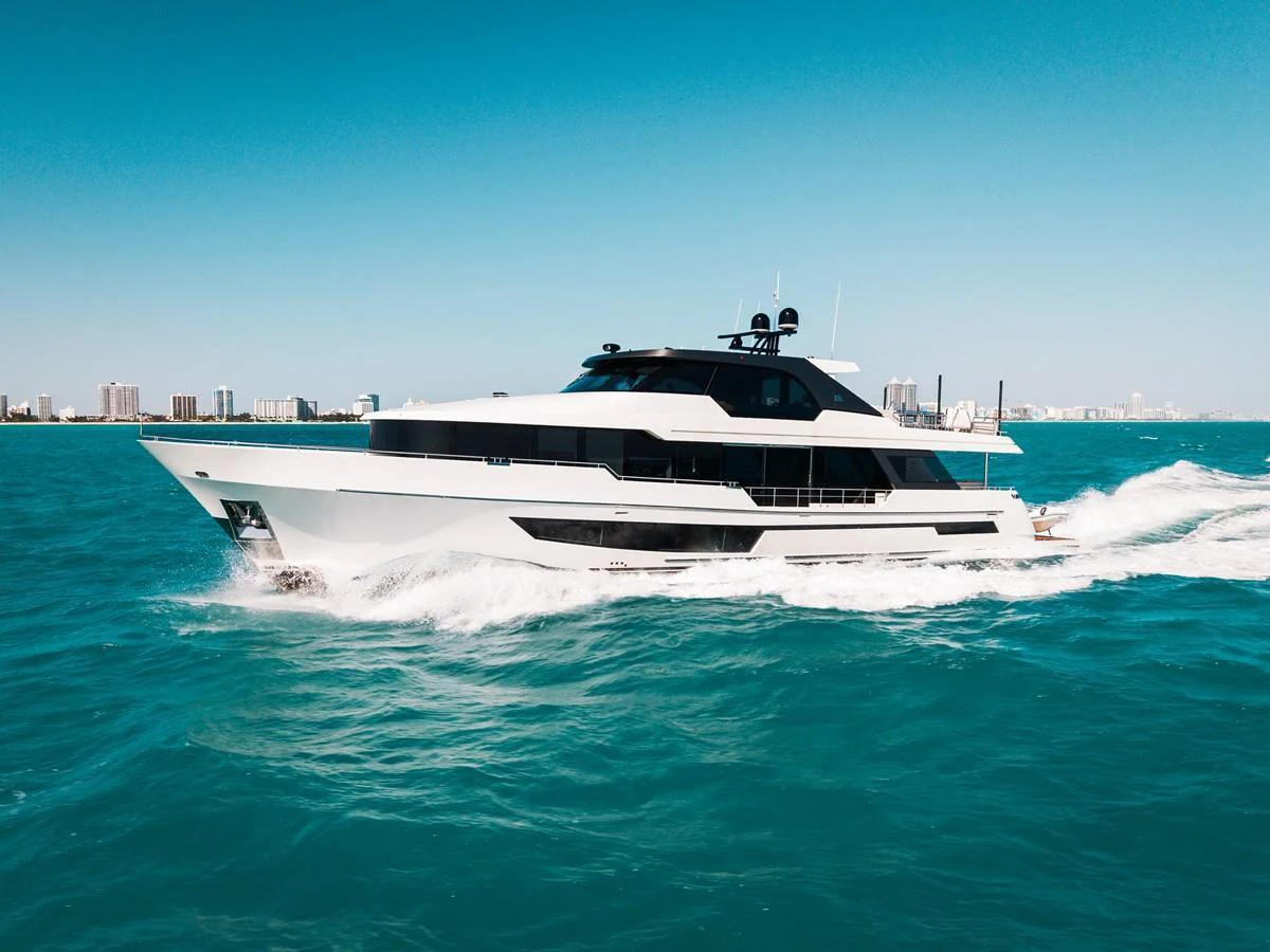A sleek and modern yacht cruises through vibrant blue waters with a cityscape in the background. aboard LA LA LA Yacht for Sale