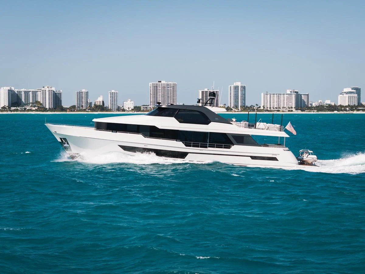 A sleek, modern yacht cruises through vibrant blue waters with a cityscape in the background. aboard LA LA LA Yacht for Sale