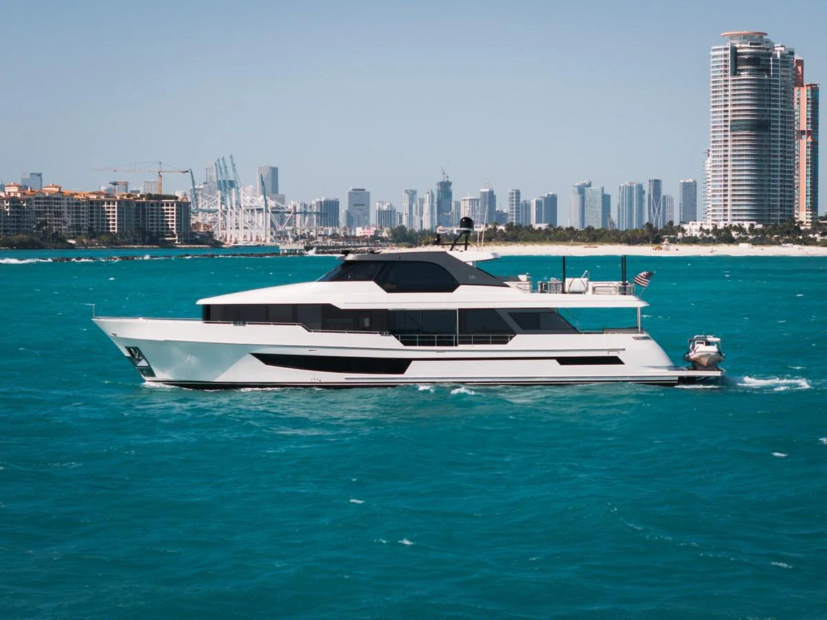 A sleek, modern yacht cruises through turquoise waters with a city skyline in the background. aboard LA LA LA Yacht for Sale