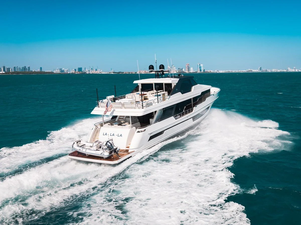 A sleek yacht cruises through vibrant blue waters with a cityscape in the background. aboard LA LA LA Yacht for Sale