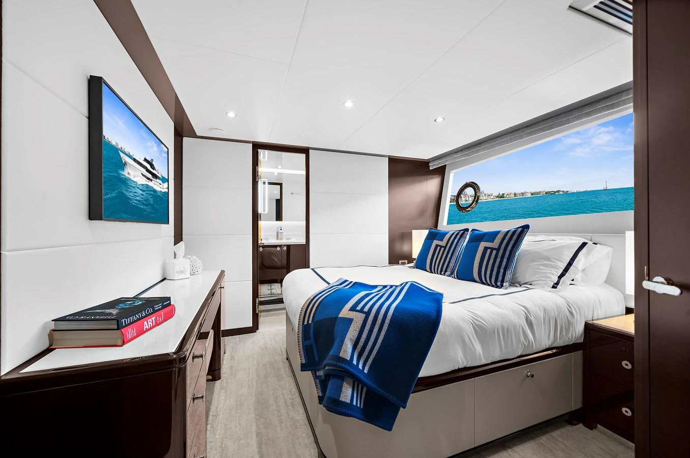 A luxurious stateroom featuring a large bed with blue and white accents, ample storage, and a view of the sea through the window. aboard LA LA LA Yacht for Sale