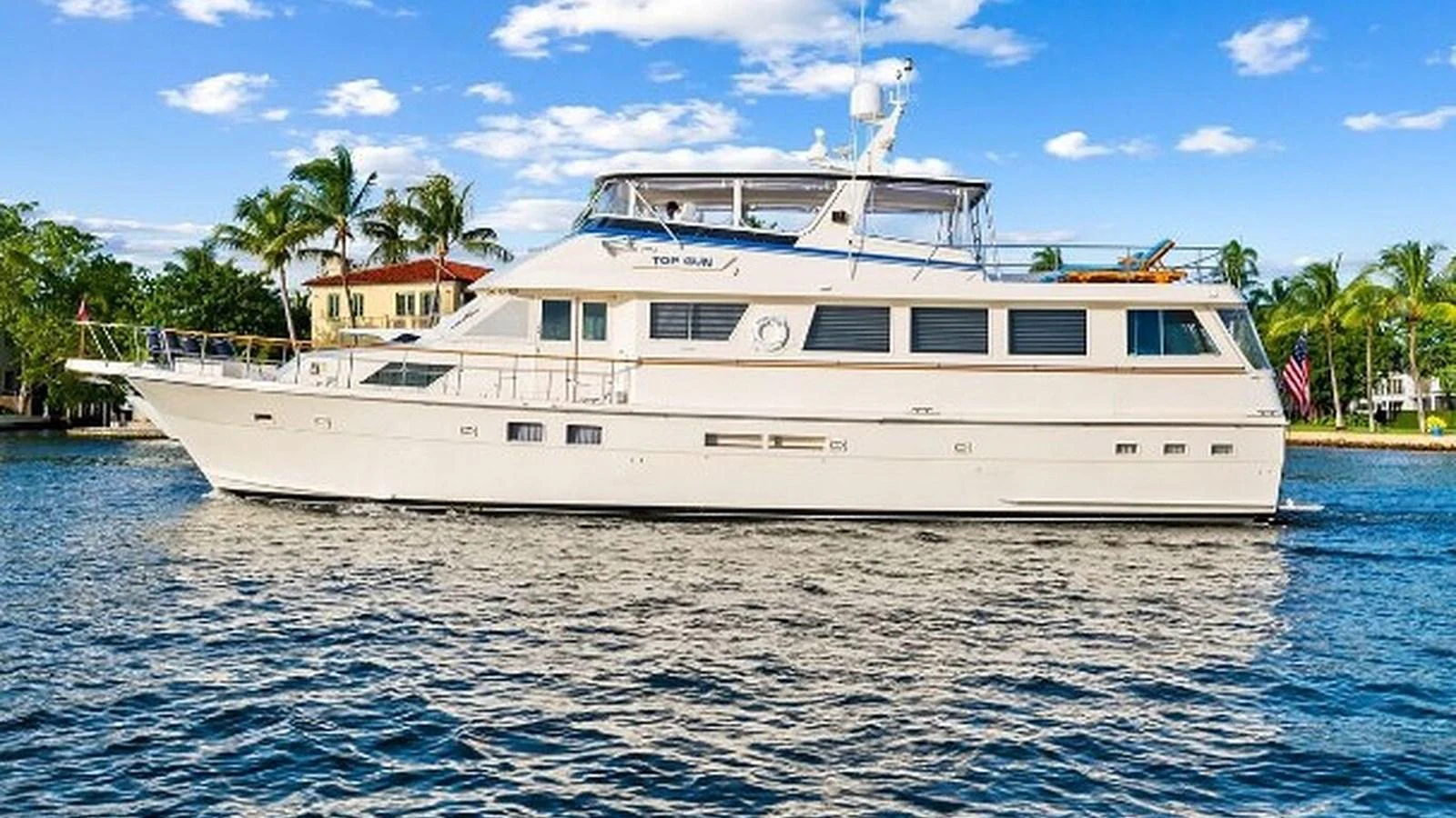 A classic motor yacht cruises gracefully on calm waters with a backdrop of lush greenery and blue skies. aboard TOP GUN Yacht for Sale