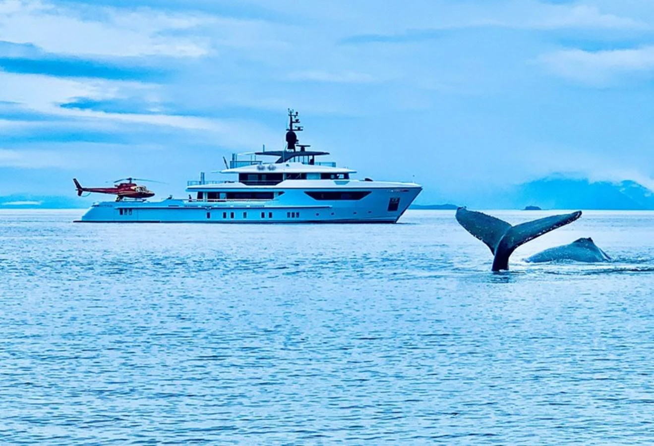 A luxurious yacht is anchored on calm waters with a helicopter on its deck and a whale tail emerging from the sea. aboard OCEAN DREAMWALKER III Yacht for Sale