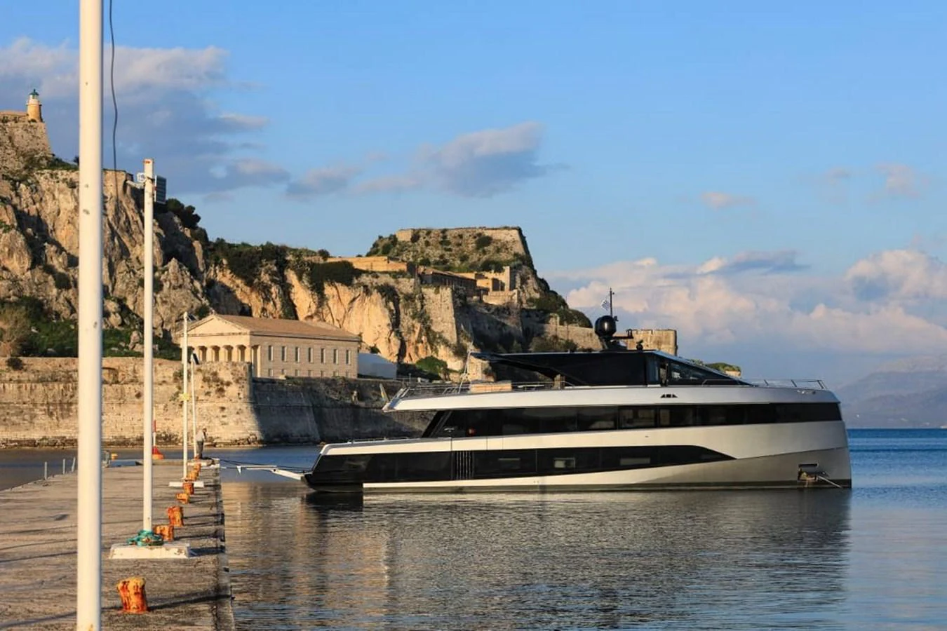 A sleek, modern yacht is docked near a historic coastal fortress, showcasing a blend of contemporary design and timeless scenery. aboard CARAT Yacht for Sale