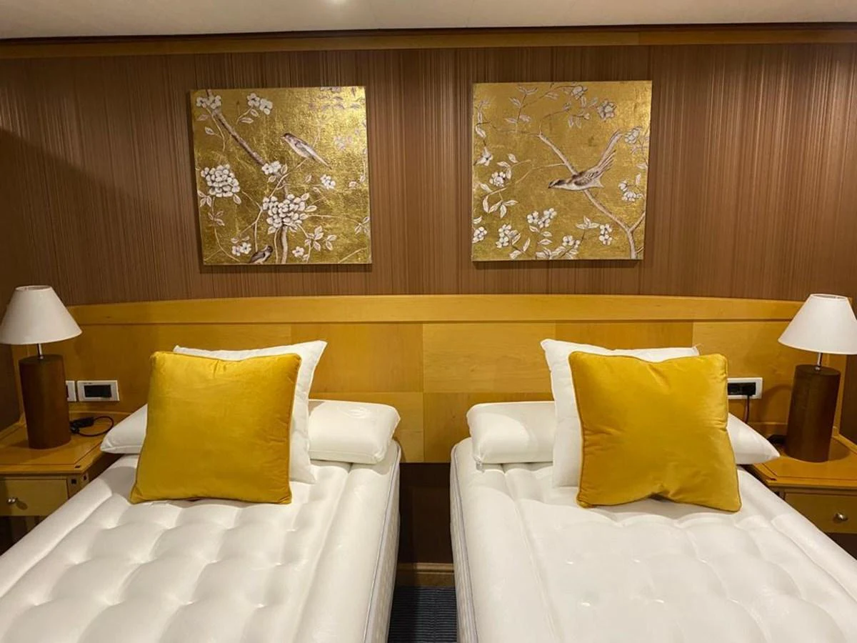 The guest stateroom features twin beds with plush white bedding and golden accent pillows, complemented by elegant wall art and warm wooden tones. aboard HARMONY II Yacht for Sale