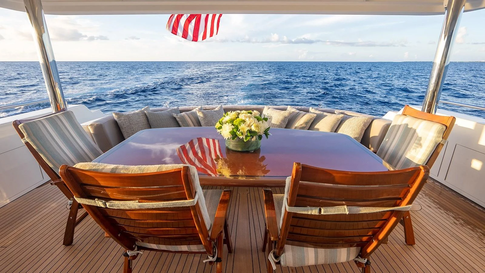 The aft deck features a polished wooden dining table surrounded by comfortable chairs, offering a perfect setting for alfresco dining with a stunning ocean view. aboard KEMOSABE Yacht for Sale
