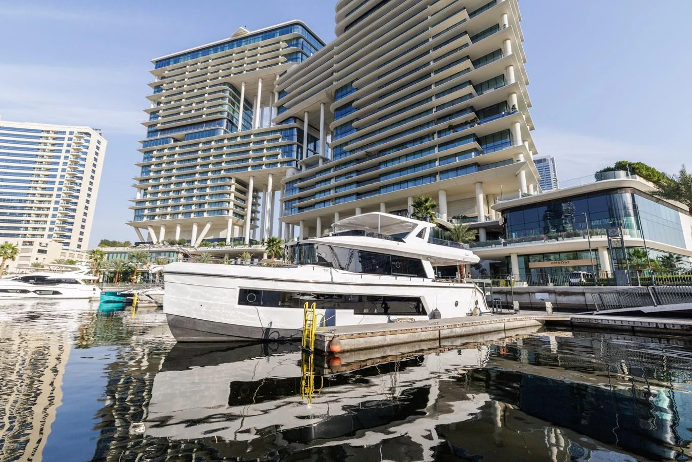 A sleek yacht is docked in a modern marina with impressive architectural buildings in the background. aboard AL SULTAN Yacht for Sale