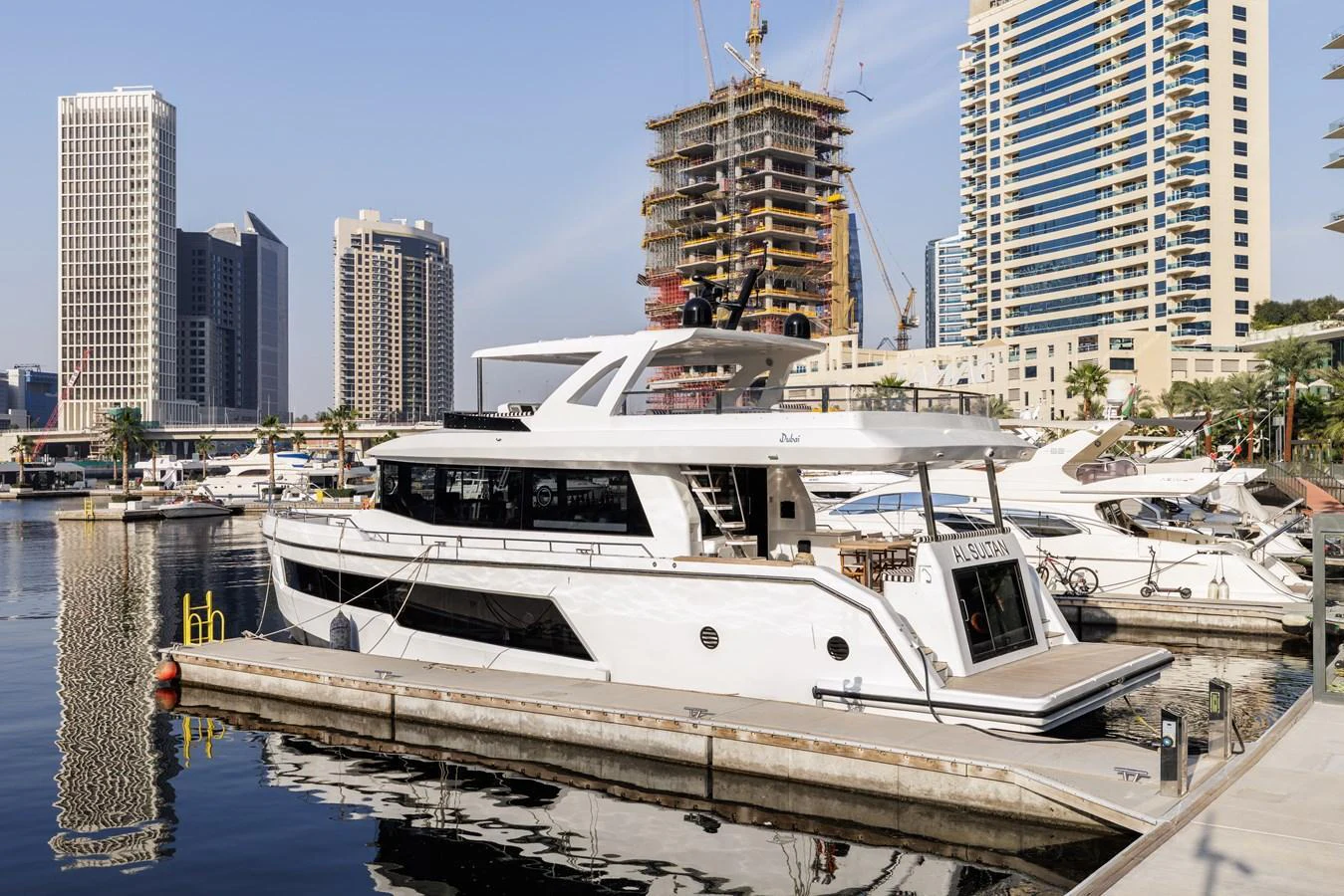 A sleek and modern yacht docked in a bustling marina with a cityscape backdrop. aboard AL SULTAN Yacht for Sale