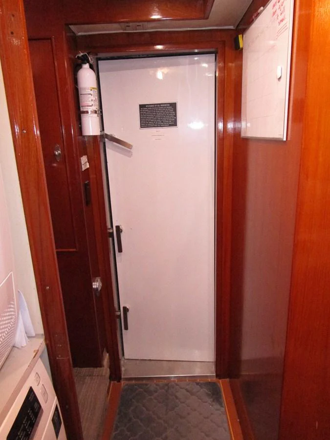 A narrow hallway with a door and wooden paneling, featuring a fire extinguisher and a whiteboard. aboard SHEPSL X Yacht for Sale