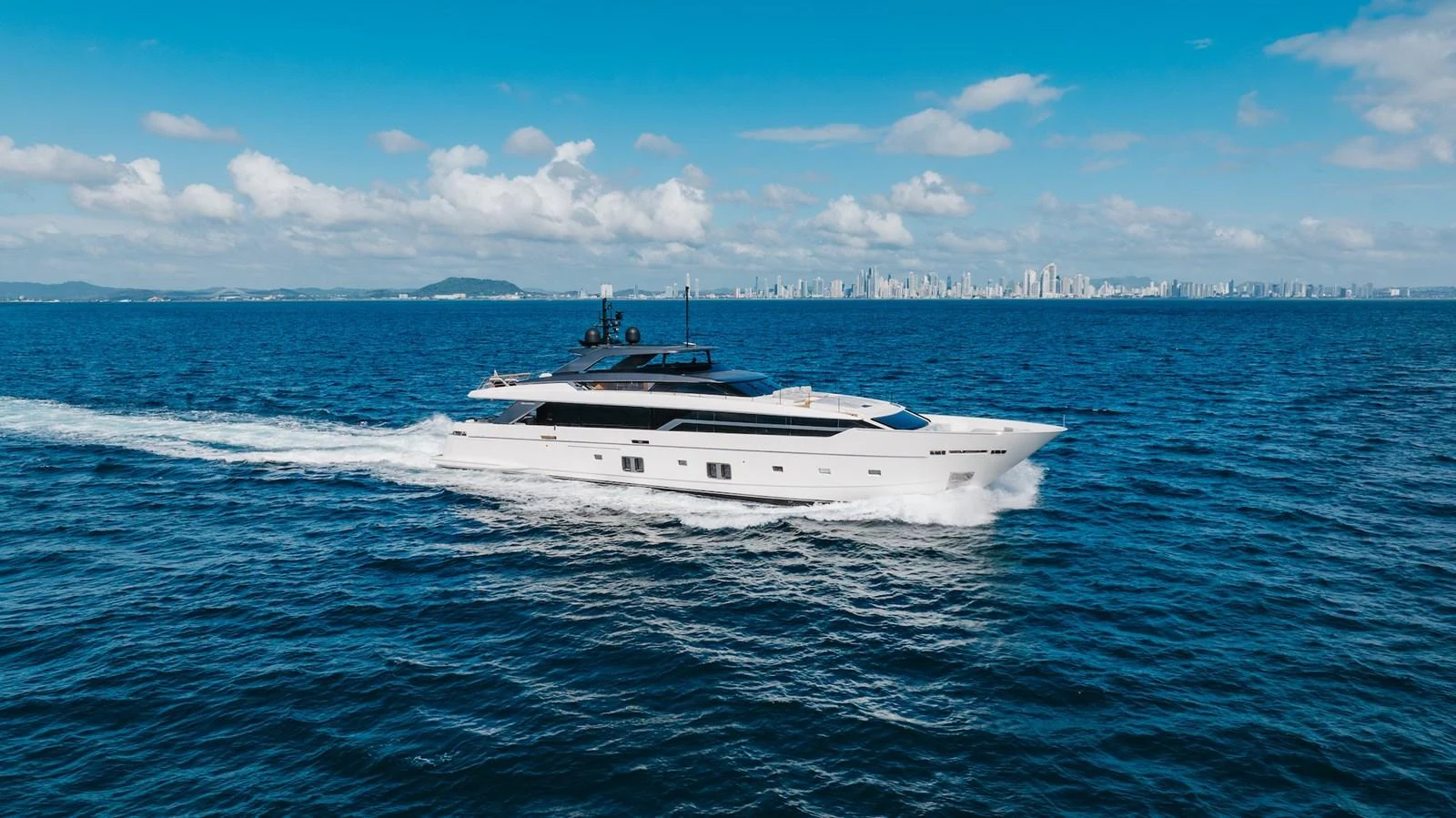 A sleek, modern yacht cruising on open water with a city skyline in the background. aboard AMBAR Yacht for Sale