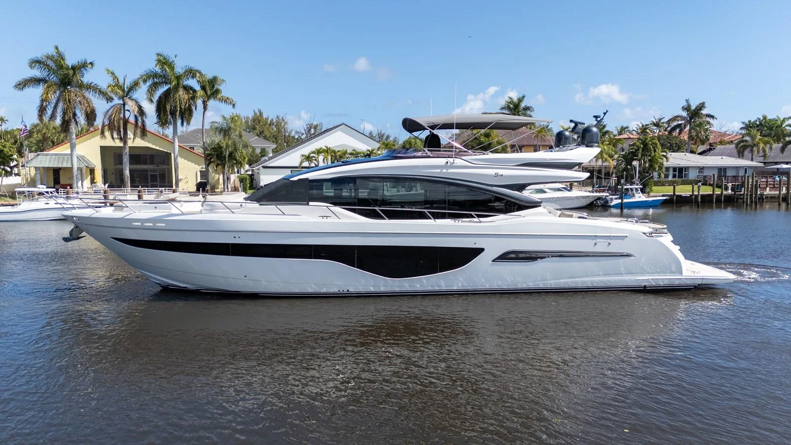 A sleek and modern yacht is cruising on calm waters with a backdrop of palm trees and waterfront homes. aboard NAUTI NATALIE Yacht for Sale