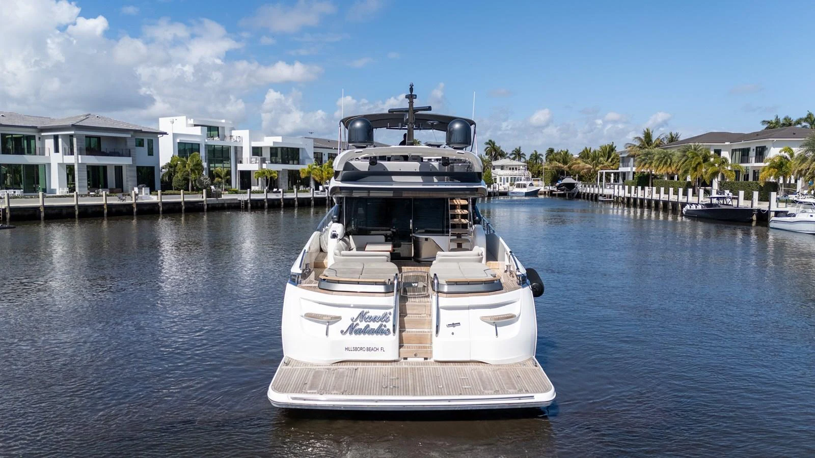 A luxurious yacht is docked in a serene marina with modern waterfront homes in the background. aboard NAUTI NATALIE Yacht for Sale