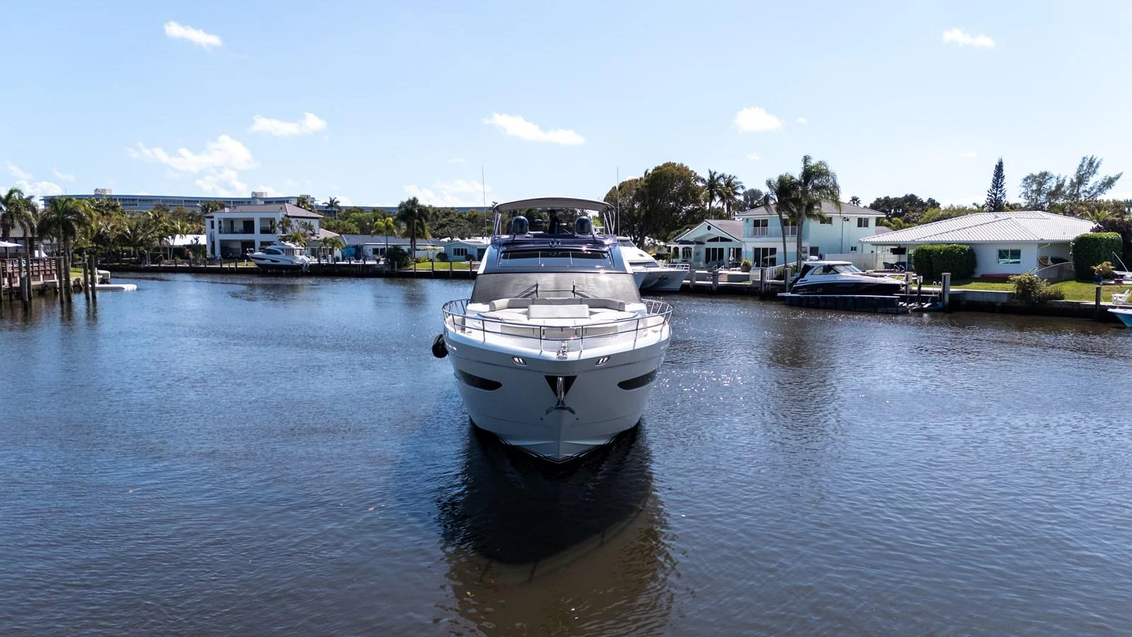 A sleek yacht is seen head-on, cruising through a calm waterway with waterfront homes in the background. aboard NAUTI NATALIE Yacht for Sale