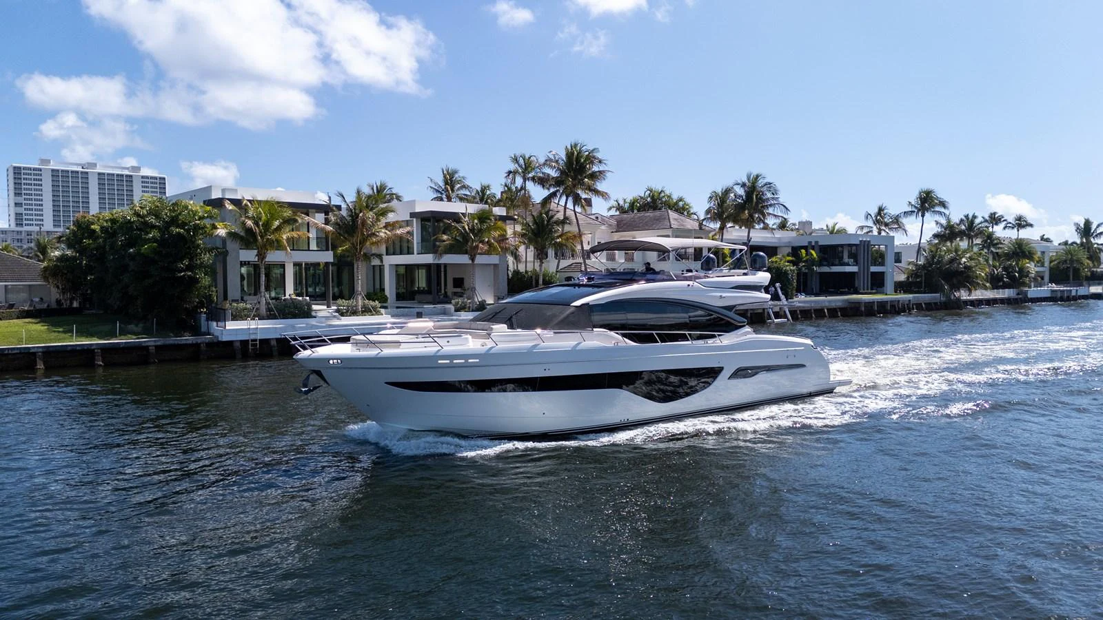 A sleek and modern yacht cruises along a waterfront lined with luxurious homes and palm trees. aboard NAUTI NATALIE Yacht for Sale