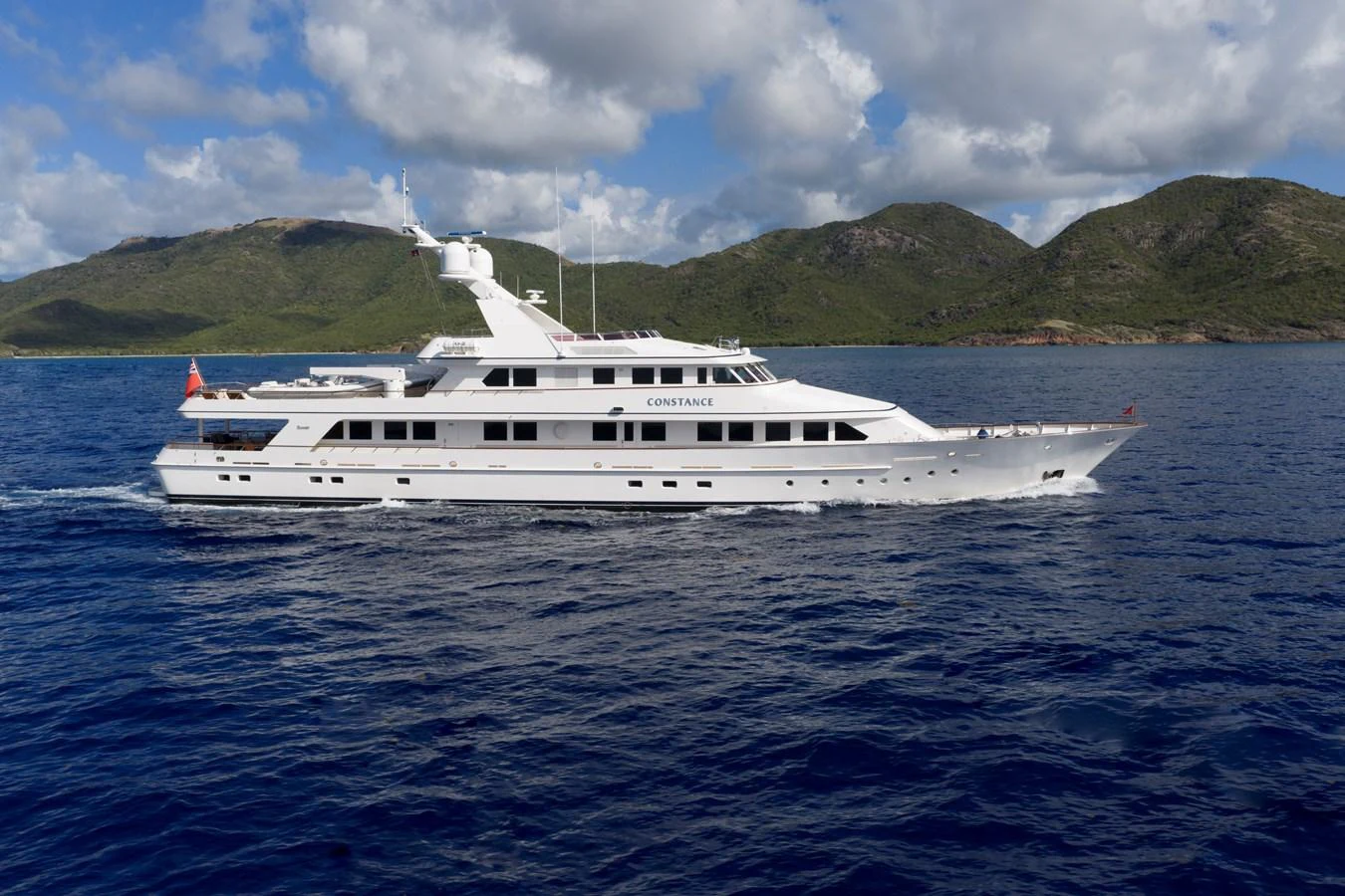 A sleek white yacht cruises through calm blue waters with a scenic mountainous backdrop. aboard CONSTANCE Yacht for Sale