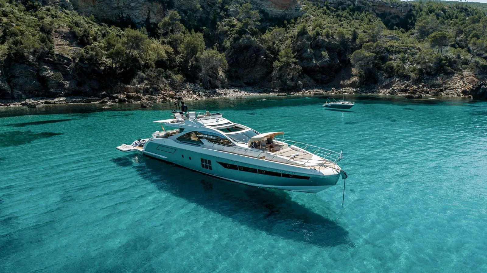 A sleek yacht is anchored in crystal-clear turquoise waters near a rocky shoreline with lush greenery. aboard GRAN ABE II Yacht for Sale