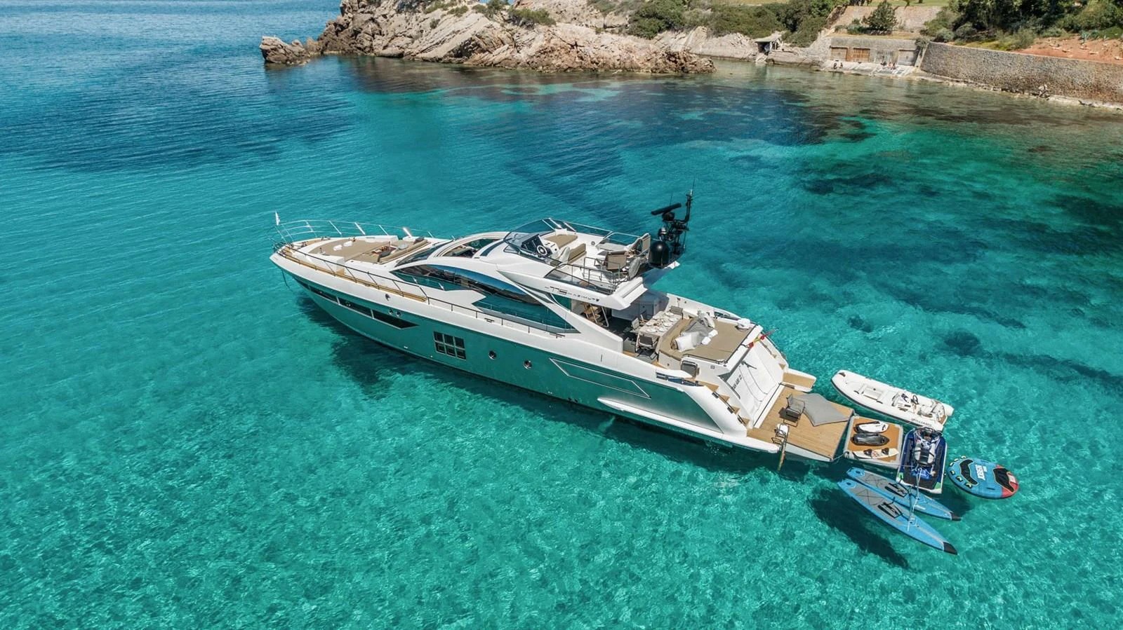 A sleek yacht is anchored in crystal-clear turquoise waters near a rocky shoreline. aboard GRAN ABE II Yacht for Sale