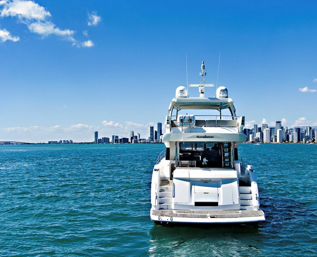 A luxurious yacht is seen from the rear, cruising on a serene body of water with a city skyline in the background. aboard SMITTEN Yacht for Sale
