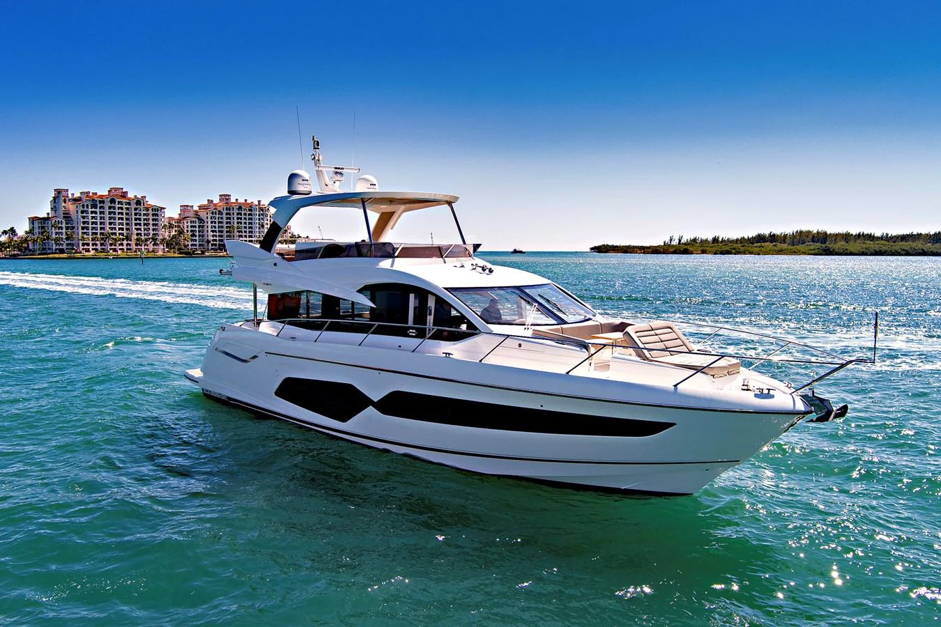 A sleek and modern yacht cruising on clear blue waters with a cityscape in the background. aboard SMITTEN Yacht for Sale