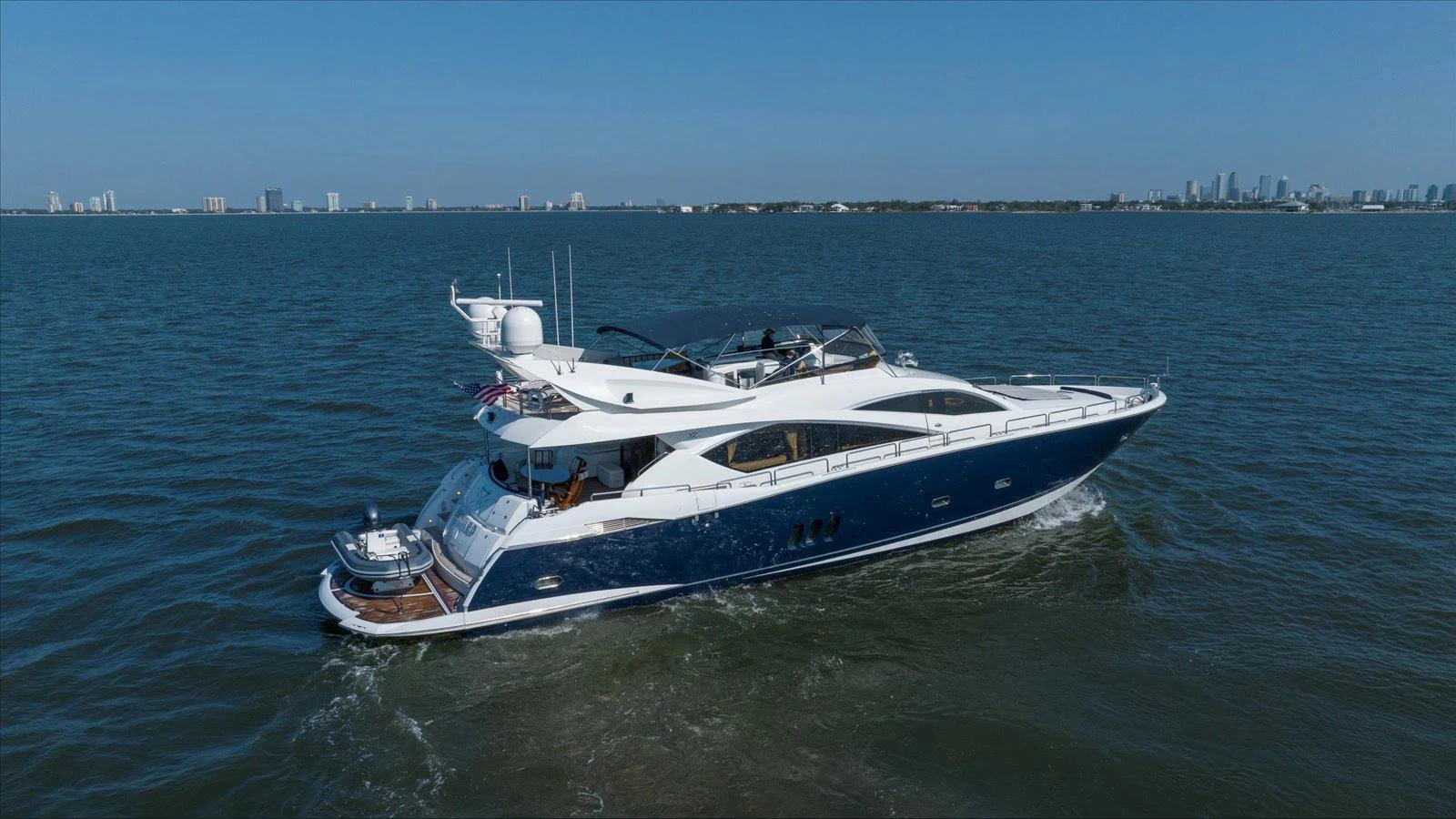 A sleek yacht cruises through calm waters with a cityscape in the background. aboard VANGUARD Yacht for Sale