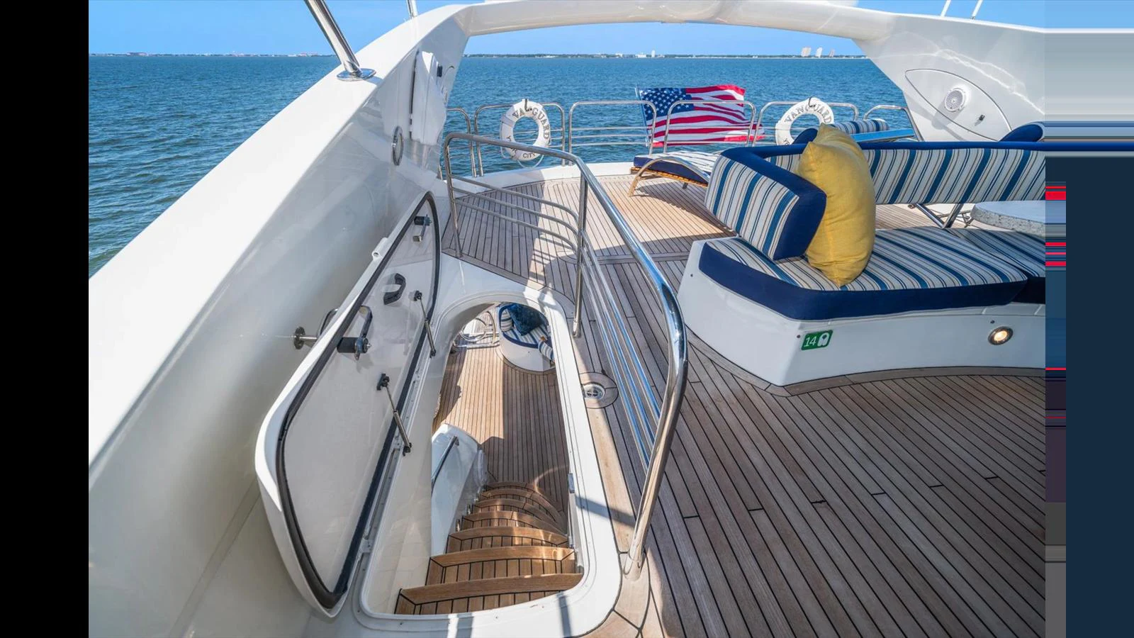 The flybridge offers a comfortable seating area with striped cushions and an American flag waving in the background. aboard VANGUARD Yacht for Sale