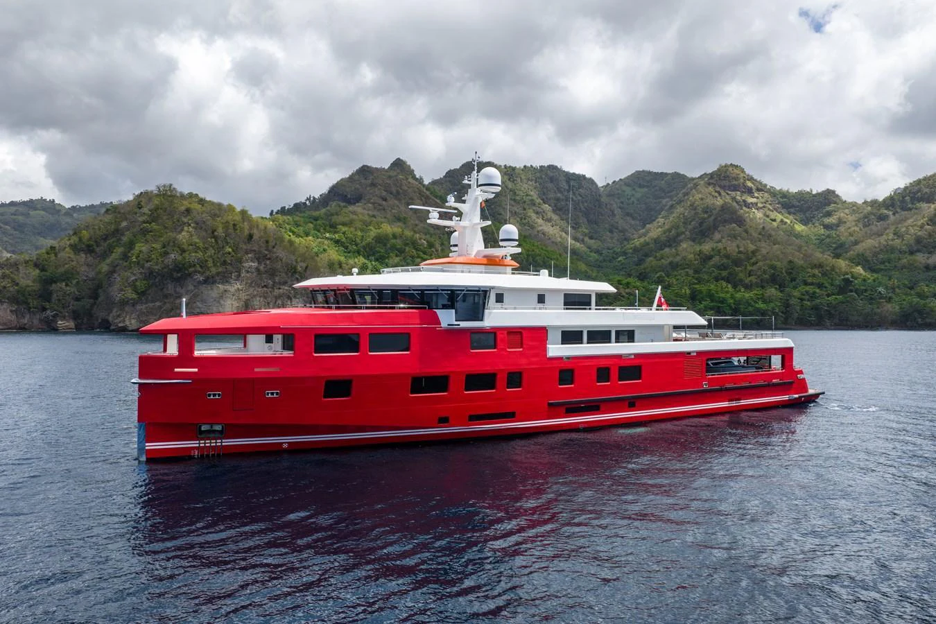 A striking red yacht cruises through serene waters with lush, mountainous scenery in the background. aboard AKULA Yacht for Sale