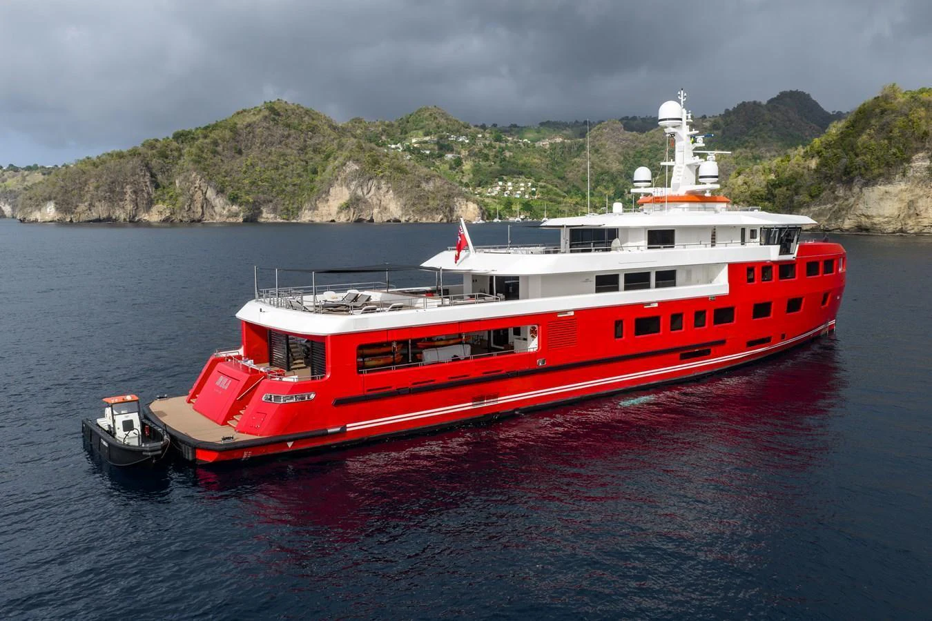 A striking red-hulled yacht is anchored in a serene bay with lush green hills in the background. aboard AKULA Yacht for Sale