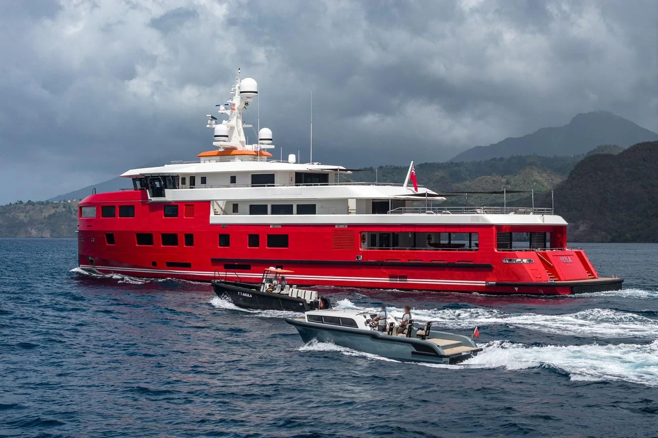 A striking red yacht cruises through the water accompanied by two smaller boats. aboard AKULA Yacht for Sale