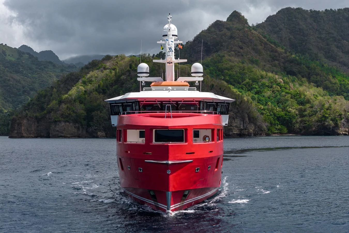 A striking red yacht cruises through calm waters with lush, mountainous scenery in the background. aboard AKULA Yacht for Sale