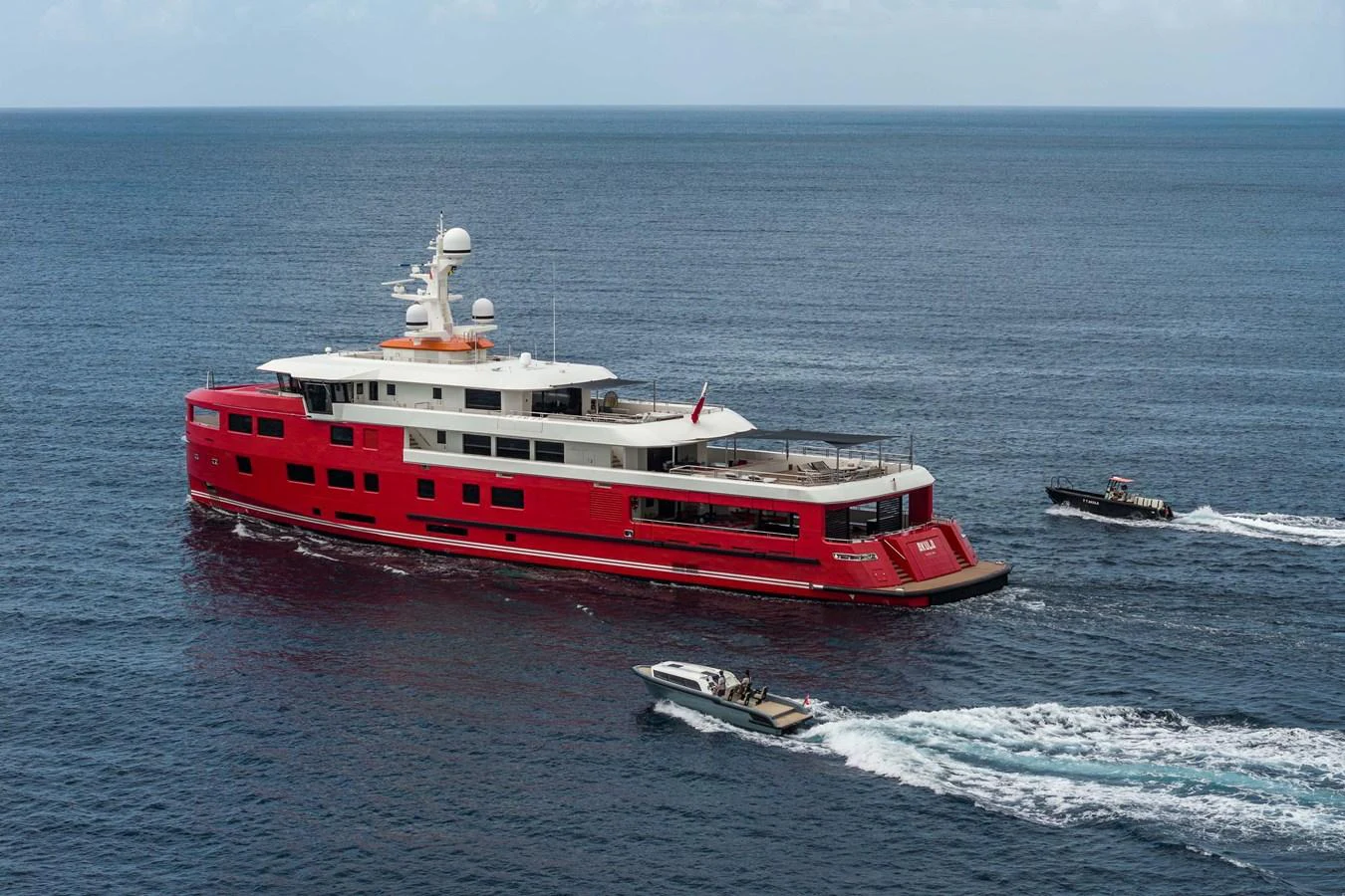 A striking red-hulled yacht cruises through calm waters, accompanied by two smaller boats. aboard AKULA Yacht for Sale