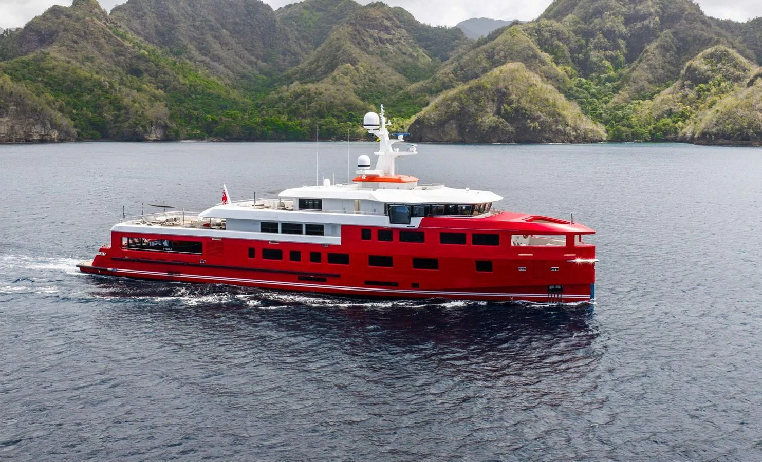 A striking red yacht cruises through serene waters with a backdrop of lush, mountainous terrain. aboard AKULA Yacht for Sale