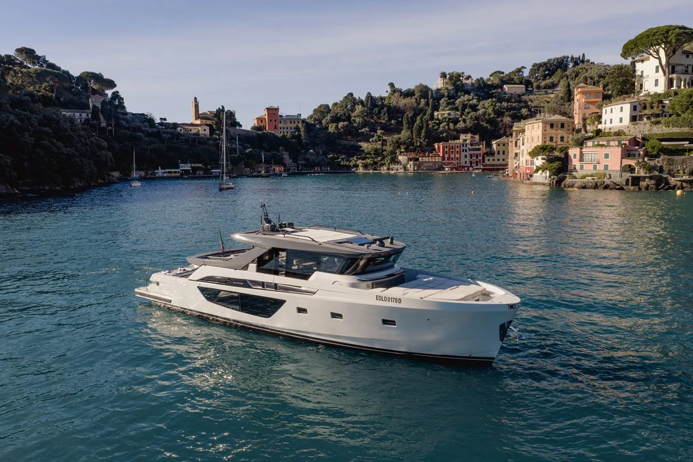 A sleek, modern yacht cruises through a picturesque coastal harbor with charming buildings in the background. aboard EOLINO Yacht for Sale