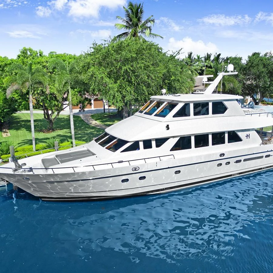 A sleek, multi-deck yacht is docked in a serene, tropical setting with lush greenery and clear blue water. aboard MILES AWEIGH Yacht for Sale